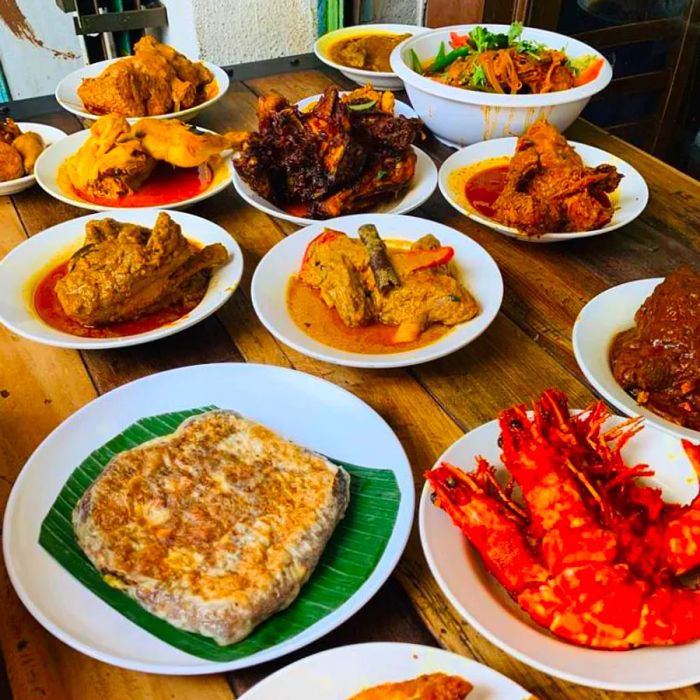 A sunlit wooden table brimming with a variety of sumptuous dishes, including large prawns, stuffed flatbread, rich curries, meaty main courses, and a generous bowl of fresh vegetable salad.