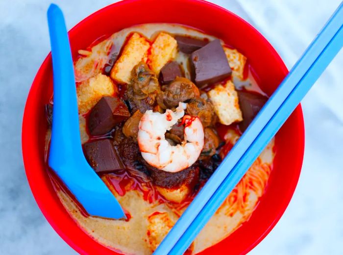 A vivid plastic bowl of curry soup, topped with a bright shrimp, sits alongside dark fried ingredients like tofu and cubes of steamed chicken blood, with a spoon and chopsticks ready for use.