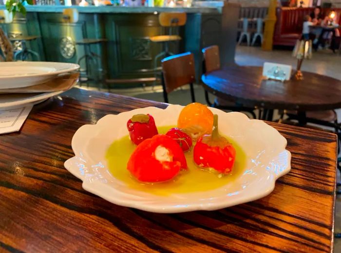 A plate of four stuffed red and orange peppers drizzled with olive oil sits on a wooden table, set against the backdrop of a cozy dining room.