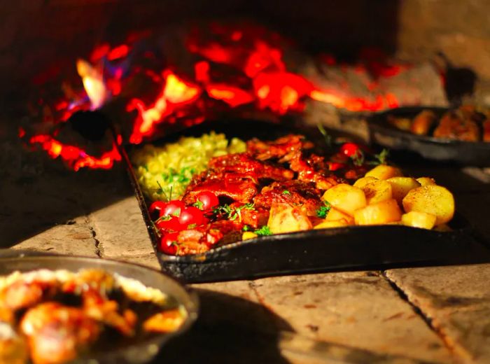 A cast iron tray filled with succulent cuts of meat, potatoes, and assorted vegetables slowly roasts over an open hearth, with other trays softly blurred in the background.