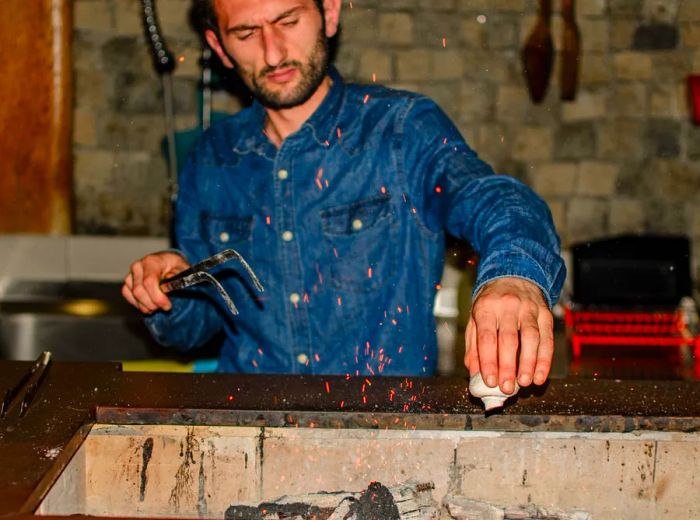 A chef carefully seasons dishes over a charcoal grill, with glowing embers rising into the air.