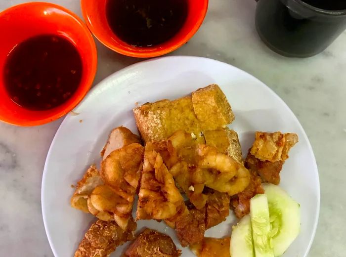 From above, a plate brimming with fried delights alongside a few slices of cucumber, two small dipping bowls of sauce, and a steaming mug of coffee or tea.