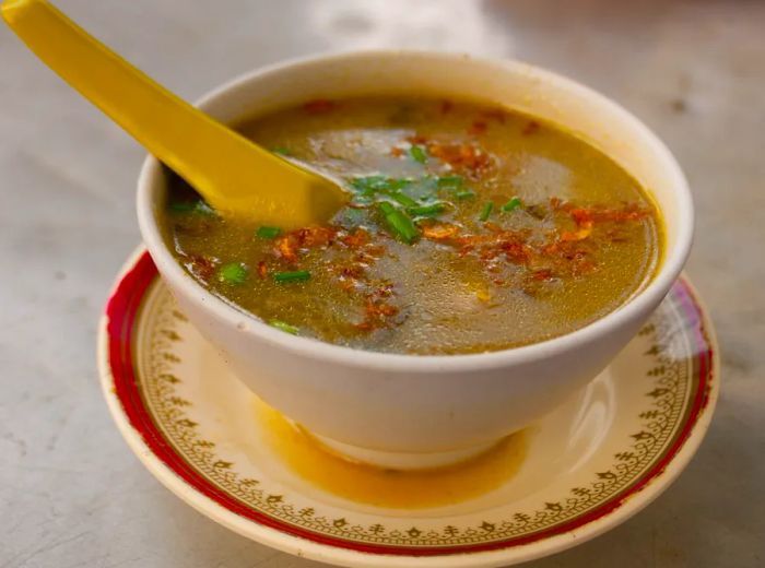 A bowl of soup placed on a saucer, featuring a thick broth sprinkled with fried shallots and chopped greens, with a large plastic spoon resting inside.