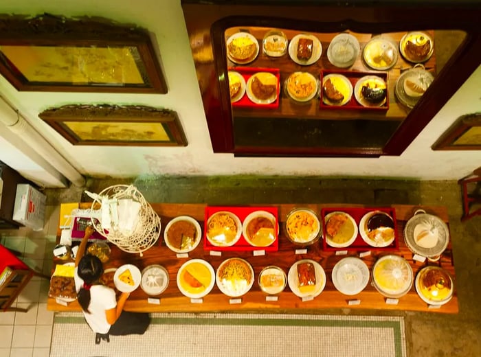 From a balcony vantage point, a server prepares a plate from a long wooden table filled with cakes and desserts, set against a wall adorned with a large mirror that reflects the treats and some obscured artworks.