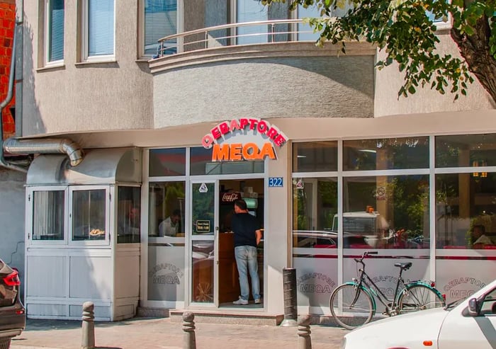 The restaurant boasts a modern exterior of gray concrete and shiny silver cladding, featuring large windows that invite diners to peek inside, along with a vibrant sign displaying the name Qebaptore Meqa.