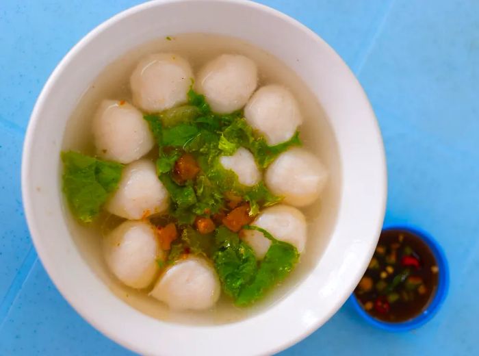 A bowl of light broth filled with fish balls and greens sits on a vibrant tile surface, accompanied by a small cup of sauce on the side.