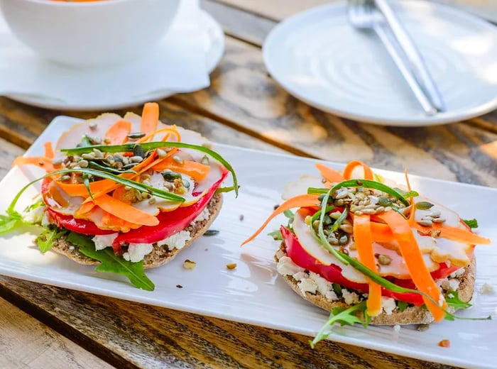 A long plate features two canapés adorned with cheese, arugula, shaved carrots, and an assortment of vegetables and pepitas.