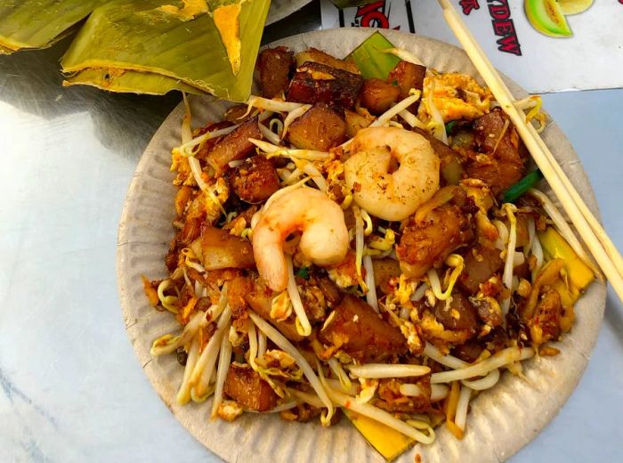 A paper plate filled with shrimp, rice cakes, and bean sprouts, chopsticks resting on the edge, alongside a banana leaf with a piece of fish curry, and a tall beverage on a plastic table featuring an illustrated menu showcasing various fruits.