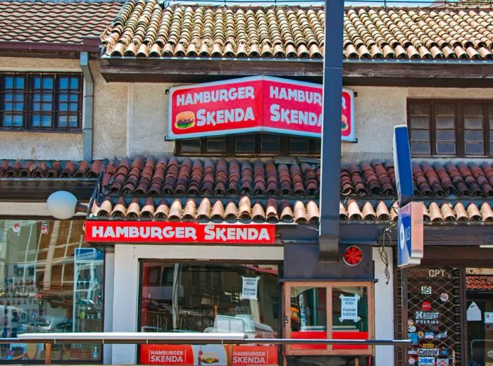 The storefront of a charming two-story building features a shingled exterior and a prominent sign for Hamburger Skenda, complete with an image of a hamburger.