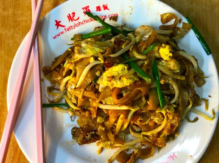 From above, a plate of stir-fried rice noodles adorned with bean sprouts, greens, and onions, with a pair of chopsticks resting on the edge of a wooden tabletop.