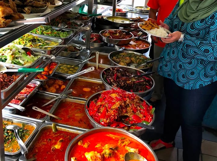 A buffet spread featuring heavy pots filled with stews and soups, smaller dishes lined up, elevated trays of vegetables, alongside rice and noodle offerings, with two customers loading their plates from the selection.