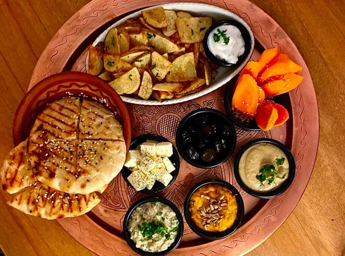 From above, a round wooden platter brimming with generous servings of potato slices and pita, accompanied by smaller bowls of dips, carrots, olives, and cheese.