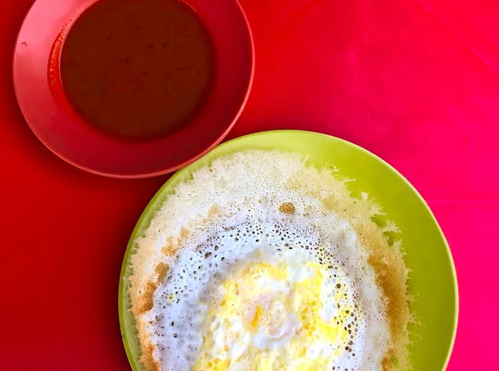 From above, a plate displays a large, sponge-like, eggy crepe next to a small bowl of sauce on a red tablecloth.