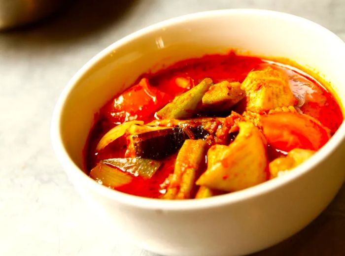 A close-up view of a bowl of soup featuring generous pieces of fish and vegetables in a thick red broth, with kitchen jars and utensils softly blurred in the background.