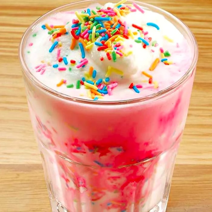 A vibrant close-up of a tall glass filled with a bright pink drink, topped with whipped cream and colorful sprinkles, set against a neutral wooden background.