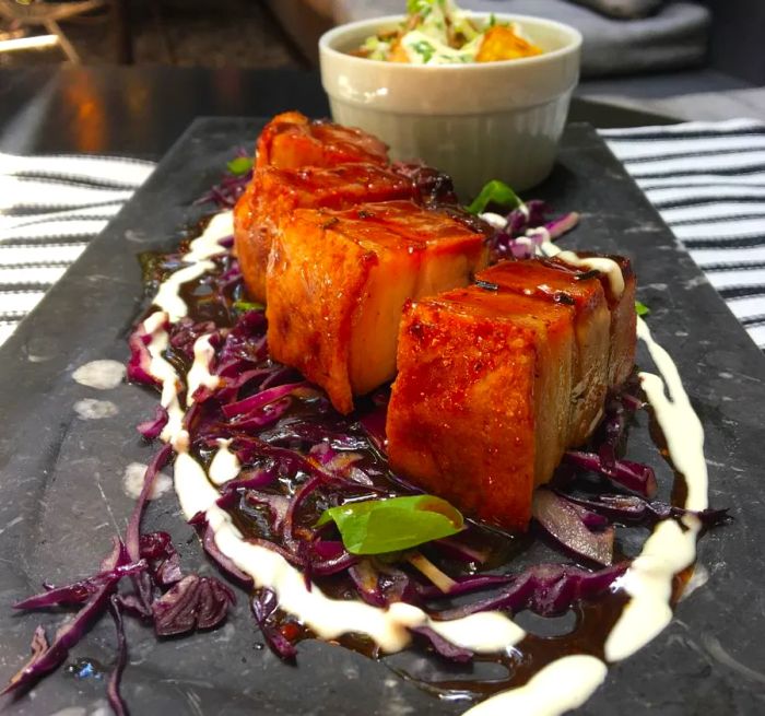 Thick slices of roasted pork belly rest on a long charcuterie board atop diced cabbage, encircled by a pool of white sauce, with a blurred bowl of dip in the background and couches in the distance.