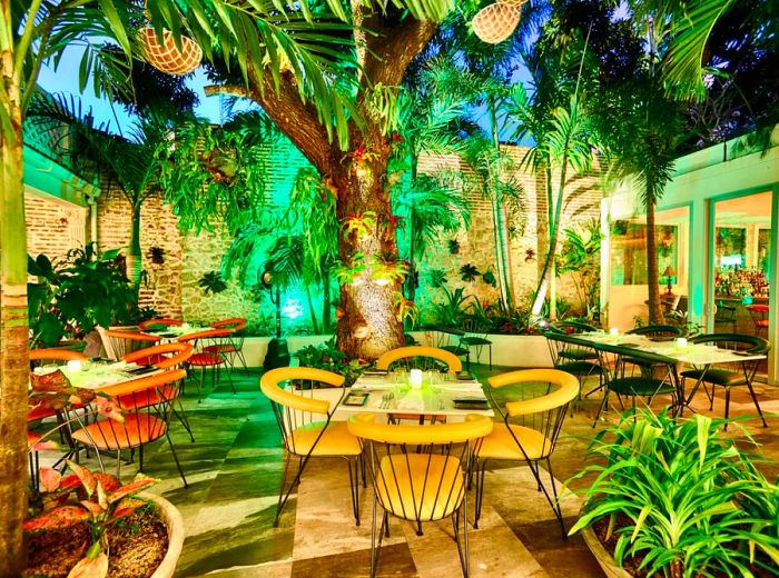 An enchanting interior courtyard featuring patio tables and chairs nestled among large trees and lush greenery, illuminated by green lights that enhance the vibrant atmosphere, with dining rooms visible on either side encircling the courtyard.