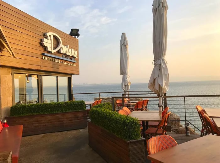 The exterior of Doniana features a prominent raised-letter sign displaying the restaurant's name, perched above a patio adorned with tables and closed umbrellas, with the sea visible just beyond the edge of the patio.