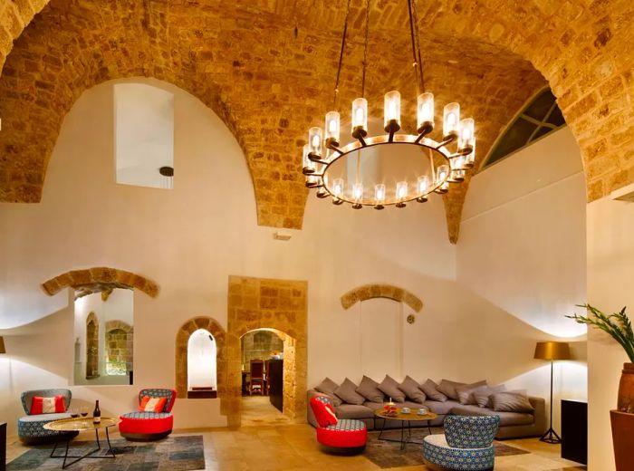 Inside a spacious hotel lounge featuring high vaulted ceilings made of exposed brick, a grand round chandelier hangs above. Cozy couches and chairs are arranged, with rooms visible through an open doorway and an interior window.