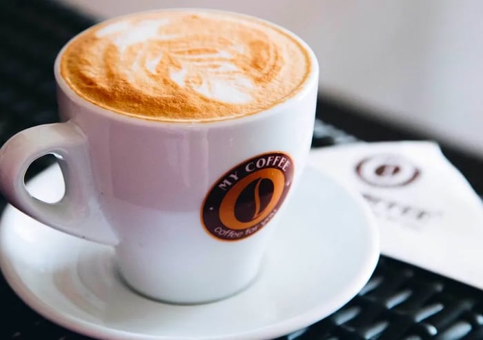 A frothy cappuccino served in a My Coffee Wings mug rests on a white saucer next to a neatly folded branded napkin.