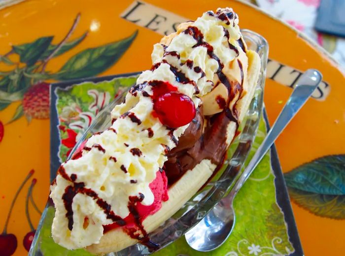 An overhead view of a classic banana split served in a long dish features sliced banana adorned with three scoops of ice cream, fluffy whipped cream, a drizzle of chocolate syrup, and a cherry on top, alongside a long ice cream spoon on a table filled with various blurred illustrations.