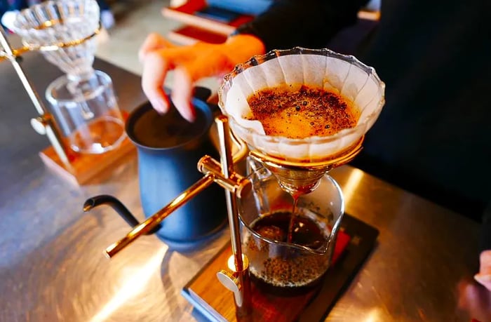 An elegant pour-over setup featuring a glass coffee beaker below, with a barista softly blurred in the background.