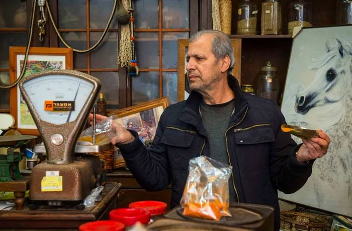 A man measures spices from a plastic bag on an antique scale, holding a scooper in his other hand, amidst a shop brimming with eclectic items