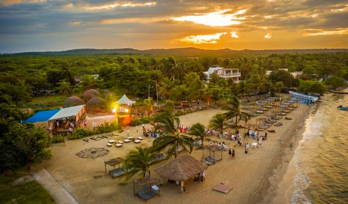 An aerial view of a sunset beach scene, showcasing trees in the distance, huts and lounge beds along the shore, with visitors below.