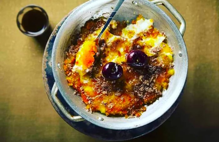 From an overhead view, a metal dish of knafeh (a pastry made from crunchy noodles) sits on a table. It has a broken brown crust with a spoon resting in the dish and two cherries perched on top, alongside a small glass of wine.