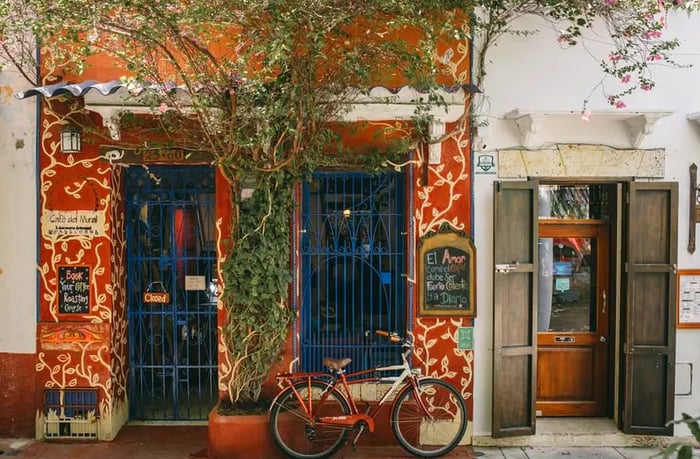 The café's exterior is adorned with vines and features blue iron gates against a red backdrop, with a bicycle parked out front and charming handwritten chalk signs advertising its offerings.