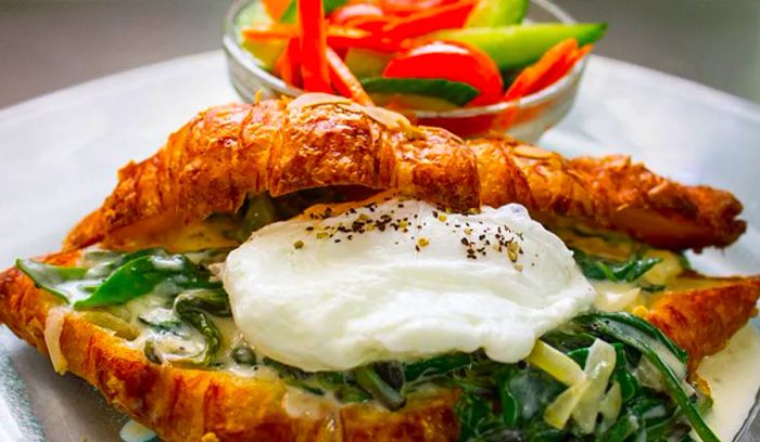 A generous scoop of fresh, gooey cheese rests inside a sliced croissant, accompanied by a bed of sautéed greens and a small dish of sliced vegetables on the plate.