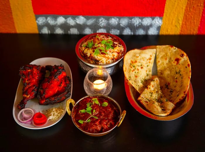 Four dishes of various shapes and materials display an assortment of Indian-Chinese delicacies, including pita, tandoori meat, rice, and curry, arranged on a dark tabletop against a vibrant wall.