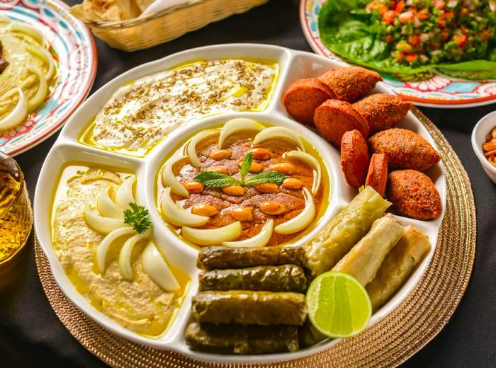 A round platter divided into sections holds various dips, falafel, and stuffed grape leaves, with additional dishes softly blurred in the background.