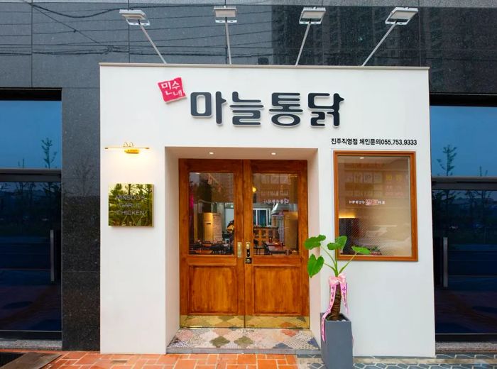 A white exterior featuring bold Korean lettering above wooden double doors, complemented by a small window with metal lights extending over the sidewalk.