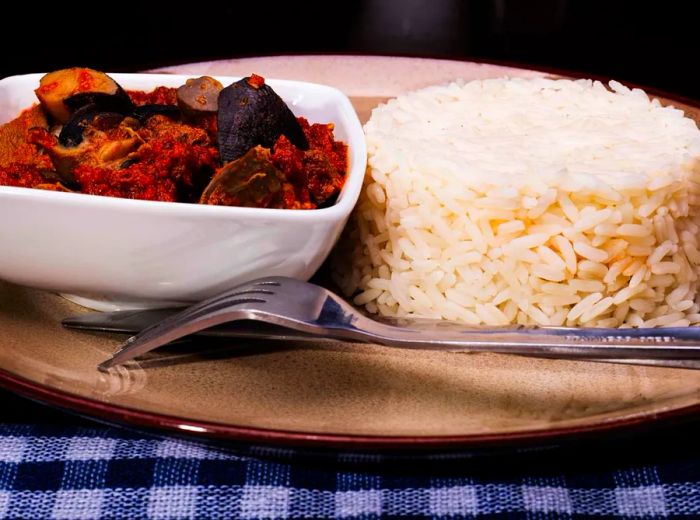 A neatly shaped mound of rice sits beside a small bowl of hearty stew, accompanied by a fork, all resting on a charming gingham tablecloth.