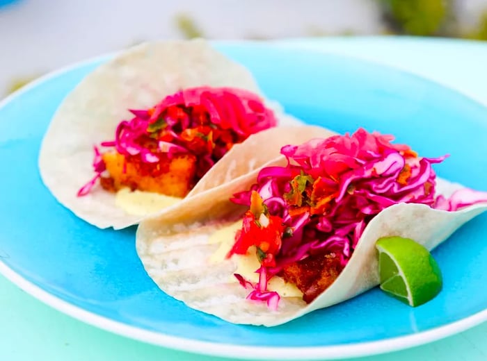 Two tacos featuring fried fish hidden beneath a mound of shredded red cabbage rest on a vibrant blue ceramic plate, accompanied by a lime wedge, on a bright formica table.