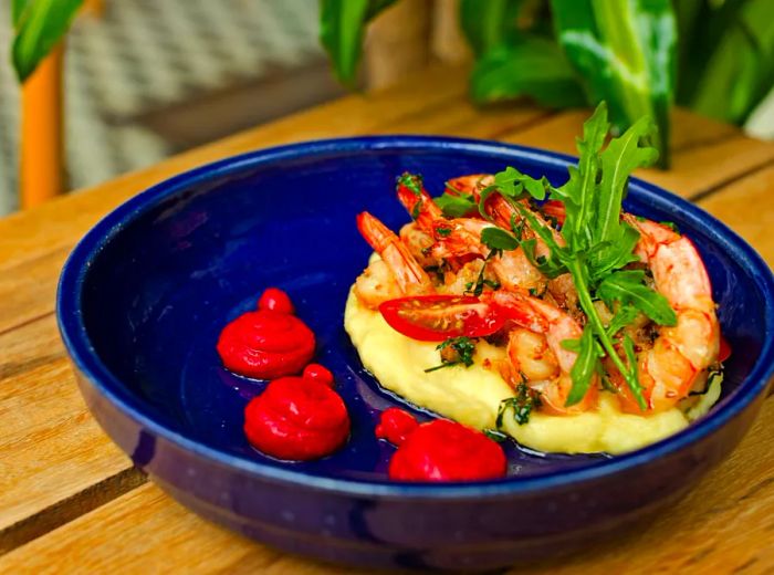 Cooked shrimp are served with tomatoes and green sprigs on a bed of mashed cassava in a ceramic bowl, accompanied by three small dollops of sauce, all set on a wooden table.