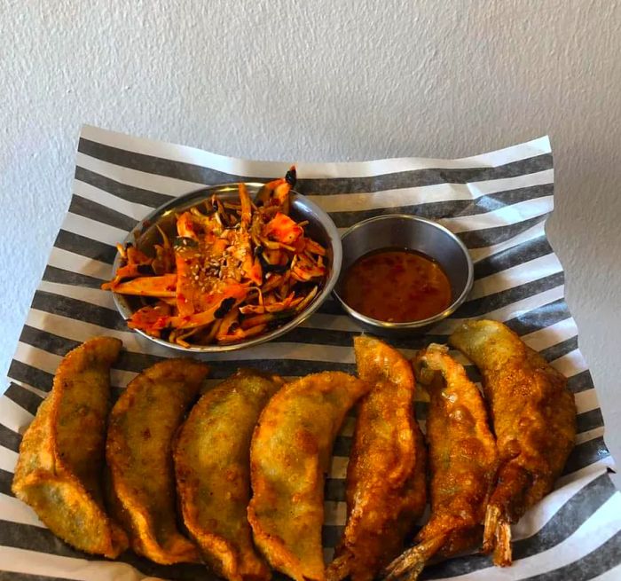 A line of dumplings arranged on striped wax paper, accompanied by a small metal dish of sauced, shredded vegetables and a cup of sauce.