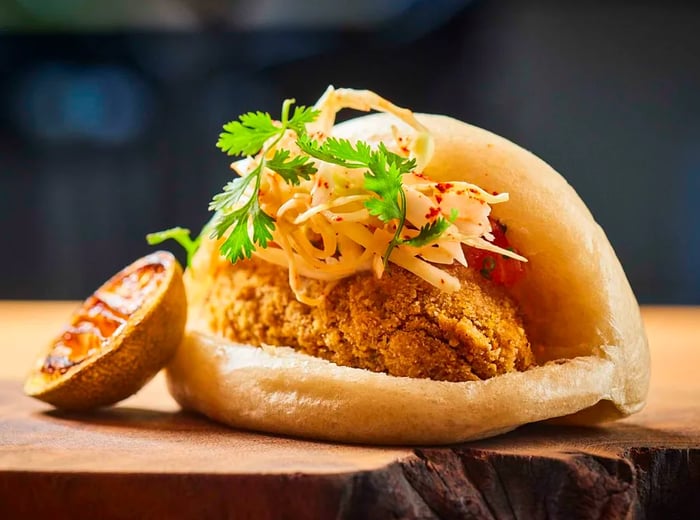 A steamed bun stuffed with fried fish, garnished with cabbage slaw and cilantro, alongside a slice of cooked lemon on a wooden surface.