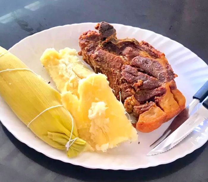 A sizable piece of chicharron rests on a white paper plate beside boiled cassava, a steaming corn husk, and silverware.