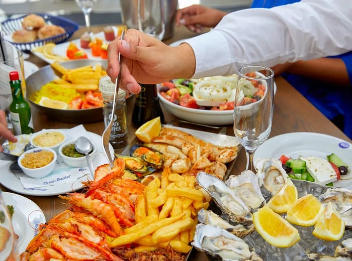 A hand reaches for a delicious morsel from a generous seafood platter, surrounded by a spread of other dishes including a tray of oysters, crispy fried seafood with French fries, Greek salad, and glasses of wine on the table.