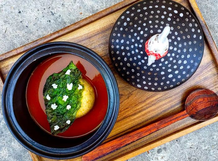 A wooden tray holds two dishes, accompanied by a long wooden spoon; one features a corn bun topped with shards of greens and delicate white flowers, while the other presents a pipe-like item on an ornate plate.