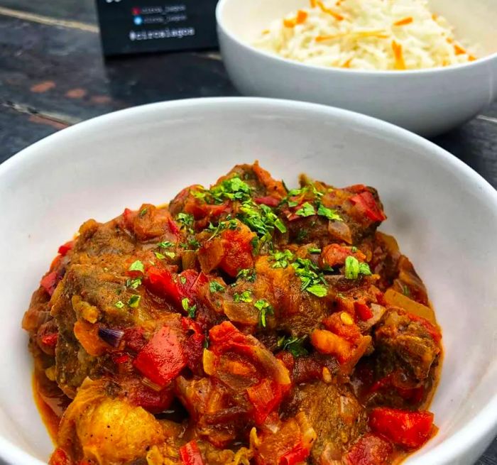 A rustic wooden table holds a white bowl filled with thick, meaty curry, showcasing tomatoes, potatoes, and tender chunks of meat, all topped with diced greens for garnish. In the background, another bowl of food is slightly out of focus.