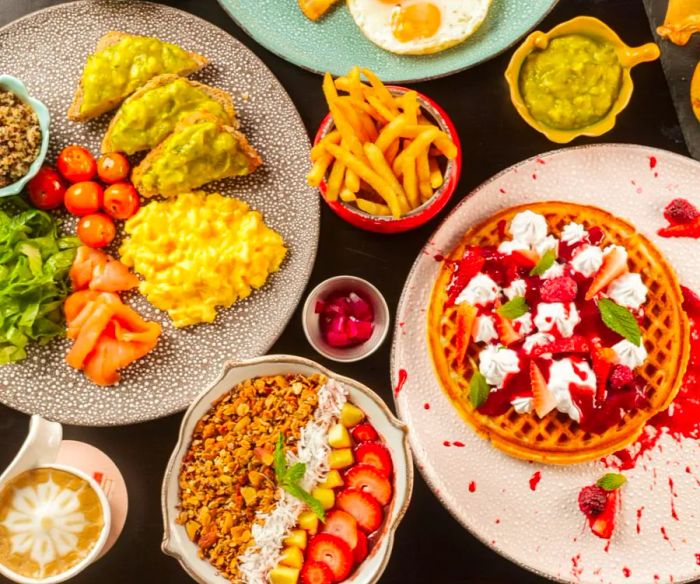 An overhead view of a delightful breakfast spread featuring a waffle adorned with whipped cream and fresh fruit slices, avocado toast topped with scrambled eggs, a bowl of granola and fruit, and crispy french fries.