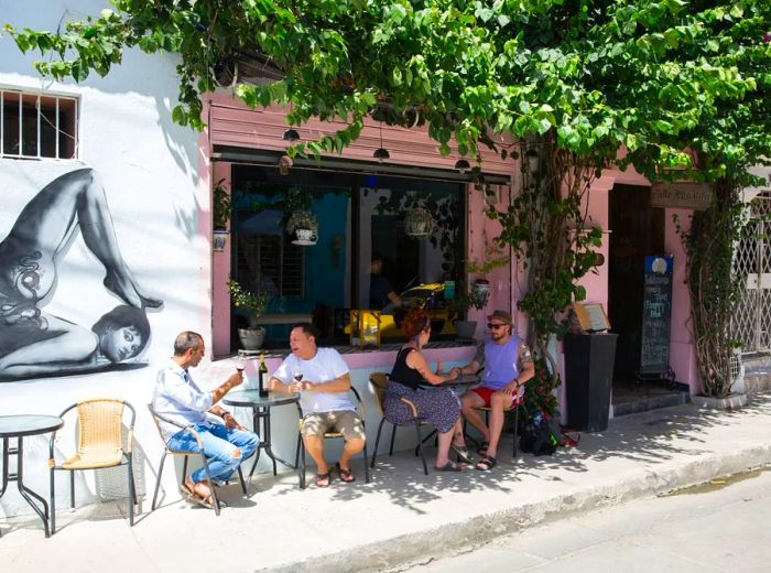 The exterior of a café features a striking grayscale illustration of a contortionist on the wall, while two small patio tables are filled with guests beneath a large window, and a door is shaded by lush foliage cascading from the floor above.