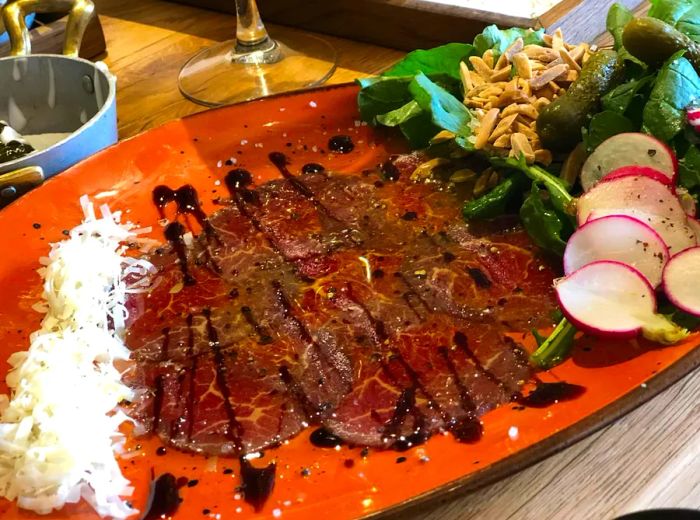 A long, vibrant plate showcases beef carpaccio, elegantly drizzled with sauce, accompanied by a salad of fresh greens, radishes, and seeds.