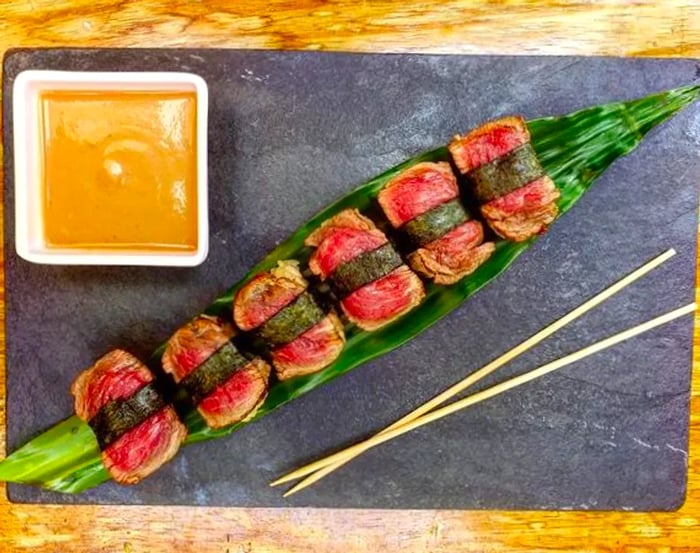 An overhead view reveals a selection of beef cuts wrapped in nori, likely resting on rice underneath, elegantly arranged on a tropical leaf atop a slate charcuterie board, accompanied by a bowl of dipping sauce and chopsticks.