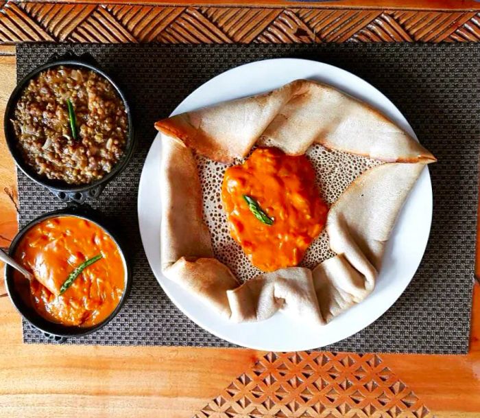 A large, folded injera flatbread rests on a white plate next to two bowls of toppings, one already placed in the center of the bread, set on a decorative wooden table with a stylish placemat.