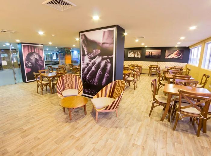 The interior of the restaurant features light wood floors, curved wooden chairs and tables, a coffee table flanked by two large-backed chairs, and a prominent central pillar adorned with grayscale images of hands and other body parts, echoing the wall decor.