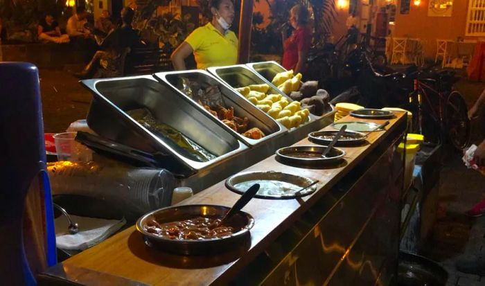 A nighttime street cart displays a row of steel containers filled with various fried snacks, positioned behind dishes of different sauces, each with spoons sticking out. A stand worker and blurred street traffic can be seen in the background.